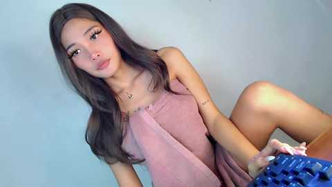 Media: A video of a young Asian woman with long, straight, dark brown hair, wearing a pink, sleeveless dress, sitting on a blue keyboard. She has a slender build, fair skin, and is indoors against a plain light gray wall.