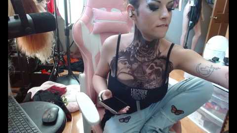 Media: Video of a tattooed woman with black and white makeup, wearing a black mesh top, sitting on a pink gaming chair in a cluttered room with gaming equipment and a computer.