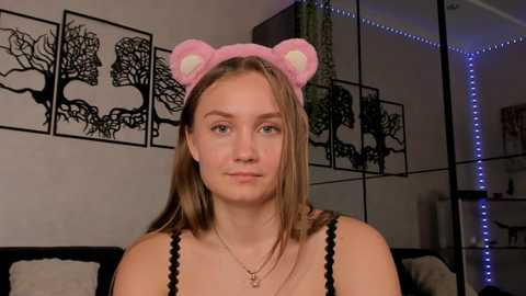 Media: Video of a young Caucasian woman with light skin, long straight brown hair, and blue eyes. She wears a pink bear headband and black lace camisole, standing indoors with tree silhouette wall art and blue LED strip lights.