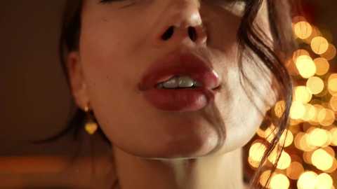 Media: Video of a close-up of a woman's face, focusing on her lips, which are slightly parted, revealing white teeth. Her skin is light, and her brown hair is slightly tousled. The background features warm, golden bokeh lights.