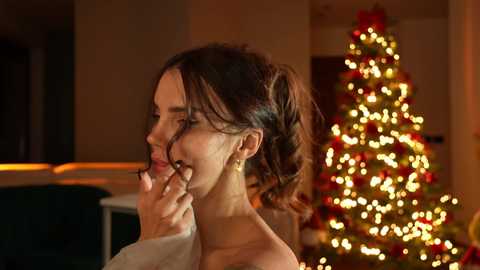 Media: Video of a smiling woman with brunette hair in a ponytail, wearing a white top, touching her nose, against a warmly lit Christmas tree with twinkling lights in the background.