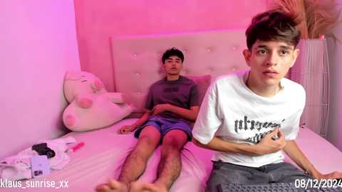 Media: Video of two young boys, one lying on a bed, the other sitting, wearing casual clothes, in a pink-lit bedroom, with toys and plushies scattered around.