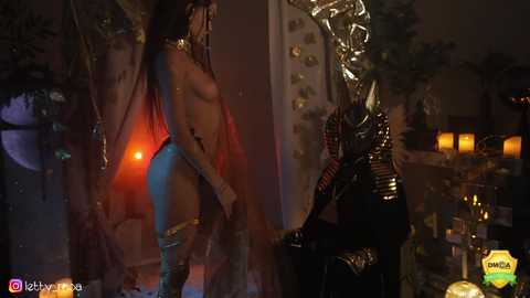 Media: A CGI screenshot of a dark, mystical scene featuring a naked woman with blue skin, long hair, and a gold headdress, standing in front of a shadowy, ornate wall.