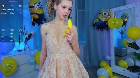 Media: Video of a young woman in a pink, sequined, strapless dress, holding a yellow banana in a playful, colorful indoor room with balloons and Minions decorations.