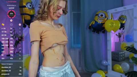 Media: Video of a slender, fair-skinned woman with curly blonde hair, wearing a cropped peach top, exposing her small breasts, and light blue pajama pants. Background features yellow Minion balloons and blue curtains.