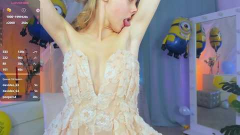 Media: Video of a blonde woman in a low-cut, lace, pale pink dress, arms raised, in a room with Minions-themed decorations, live stream overlay on screen.