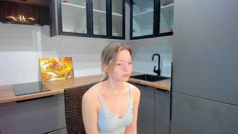 Media: Video of a young woman with light skin and blonde hair, wearing a light blue tank top, sitting in a modern kitchen with grey cabinets, a wooden countertop, and a colorful painting on the wall.