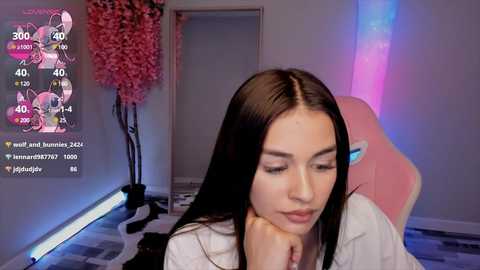 Media: Video of a young woman with long black hair, wearing a white shirt, sitting on a pink gaming chair in a modern room with a pink tree and digital stats on the wall.