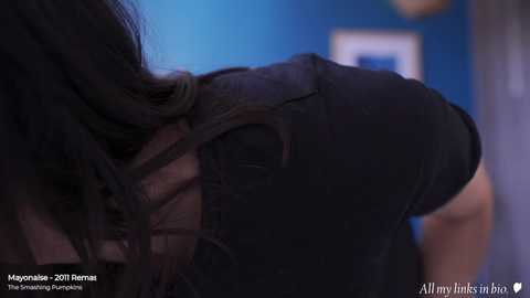 Media: Video of a person with long dark hair, wearing a black jacket, leaning against a blue wall. The background is blurred.