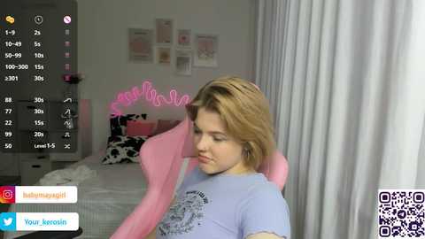 A video of a young woman with light skin and shoulder-length blonde hair, wearing a light blue graphic t-shirt, sitting on a pink gaming chair in a pastel-themed bedroom.