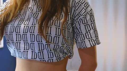 Media: Video of a woman with light skin and long brown hair, wearing a white crop top with black Roman numeral pattern, standing against a blue and white background.