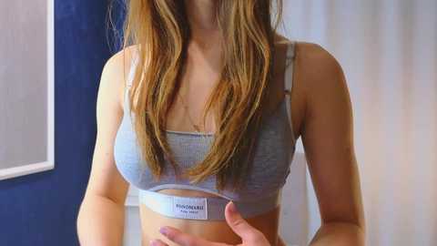 Media: Video of a young woman with long, straight brown hair, wearing a light blue sports bra with a white brand name tag, standing indoors with a blue wall and white curtains in the background.