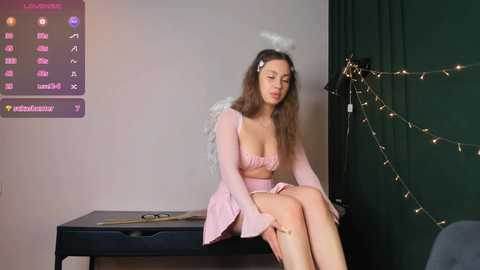 Media: Video of a young woman with long brown hair and fair skin, wearing a pink, angel-themed outfit, sitting on a black table. She has a small white halo and wings. Background includes a green curtain with fairy lights and a screen displaying text.