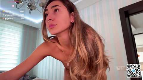 Media: Video of a topless, fair-skinned woman with long, wavy, blonde hair, wearing a nose ring, in a brightly lit room with striped wallpaper and blinds.