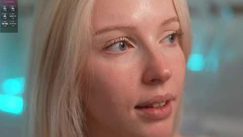 Video of a close-up of a fair-skinned woman with straight blonde hair, blue eyes, and slightly parted lips, gazing slightly to the left. The background is blurred, with a hint of blue lighting.