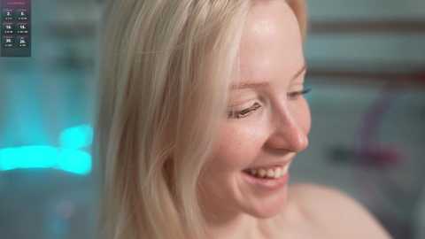 A video of a smiling, fair-skinned woman with long blonde hair, captured from the shoulders up. She is indoors with blurred background lights.