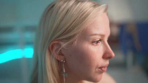 Video of a blonde Caucasian woman with straight hair, light skin, and a contemplative expression. She wears dangling silver earrings. Background is blurred, suggesting an indoor setting with soft lighting.