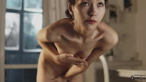 Media: A video of a slender, nude Asian woman with small breasts, leaning forward, her hands covering her chest, standing in a modern, softly lit kitchen with large windows.