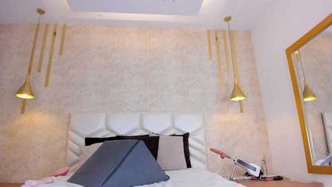 Media: A video of a modern, well-lit bedroom with beige textured wallpaper, a white tufted headboard, two pendant lamps, a mirror, and a grey mattress.