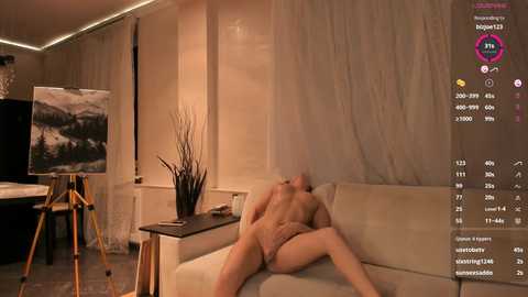 Media: Video of a nude man with fair skin, light brown hair, and medium build, lying on a beige couch in a modern, dimly lit room.