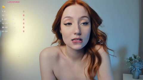 Media: A video of a fair-skinned, red-haired woman with a slender physique, wearing only a nose ring. She has a soft, sensual expression, with a hint of a smile. Background features a plain beige wall and a vase with flowers.