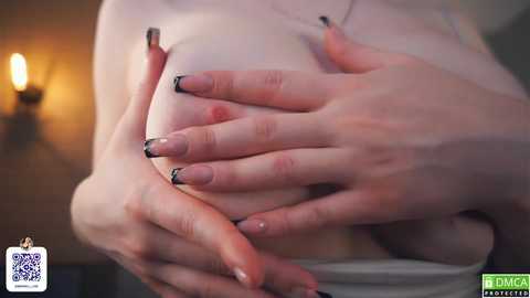 Media: Video of a pale-skinned woman's hands gently cupping her bare breast, featuring black-and-white French-tipped nails. Background shows a dimly lit room with a warm, soft glow. QR code in lower left corner.