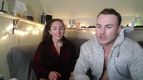 Media: Video of a young couple seated on a couch in a cozy, dimly lit living room. The woman, with long brown hair, wears a red plaid shirt. The man, with short brown hair, dons a gray hoodie. Shelves and string lights adorn the background.