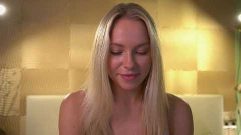 Media: Video of a blonde woman with straight hair, fair skin, and a closed expression, standing in a bathroom with a yellow tiled wall and a white sink in the background.