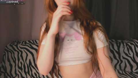 Media: Video of a fair-skinned woman with long, straight red hair, wearing a white crop top with a pink bunny logo, and zebra-patterned shorts, sitting on a couch.