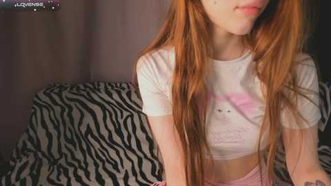 Video of a young woman with long red hair, wearing a white crop top and pink pants, sitting on a zebra-patterned couch, indoors.