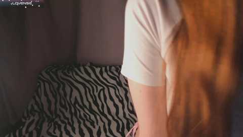 Video of a woman's upper body in a white T-shirt, leaning against a zebra-print couch in a dimly lit room.