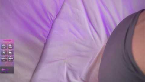 A video of a person lying on a purple bedspread, with a gray bra visible on the right side. The background includes a digital display with health metrics, such as heart rate and oxygen levels.