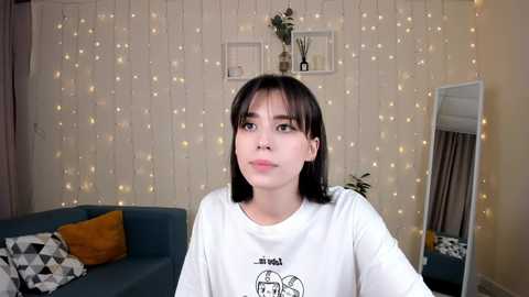 Media: Video of a young Asian woman with straight black hair and bangs, wearing a white T-shirt with a cartoon character, in a cozy living room with string lights, a blue couch, and plants.