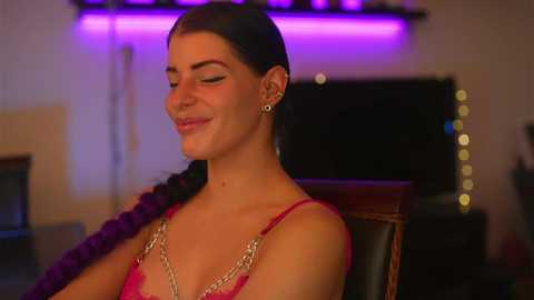 Media: Video of a smiling, fair-skinned woman with long dark hair in a pink lace bra, wearing gold chain earrings, seated in a dimly lit room with purple and white ambient lighting.