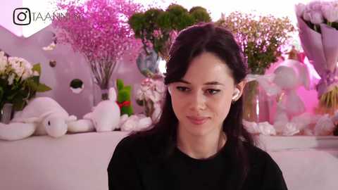 Media: A video of a young woman with dark hair and fair skin, wearing a black top, standing in a whimsical, pink-themed room with plush toys and floral decorations.