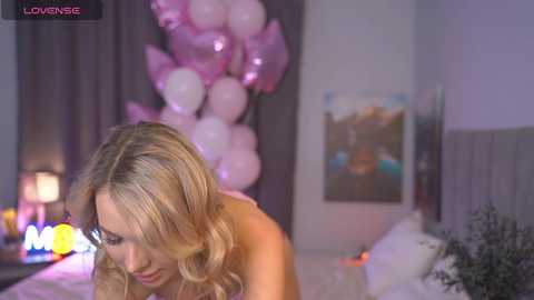 Media: Video of a blonde woman with wavy hair, topless, leaning forward, in a dimly lit bedroom with pastel balloons and a bed in the background.