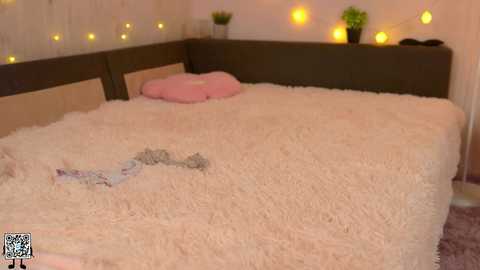 Media: Video of a cozy, softly lit bedroom with a plush, cream-colored shag rug on the bed, a pink pillow, and a green plant on a dark wooden headboard.