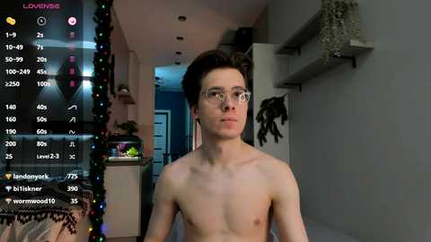 Media: Video of a shirtless young man with pale skin, glasses, and short dark hair, standing in a dimly lit room with a live stream overlay.