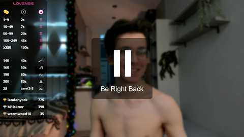 Media: A blurry video of a young man with short dark hair, wearing glasses, and no shirt, standing in a dimly lit room with festive decorations. \"Be Right Back\" overlay.