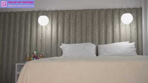 Media: Video of a modern bedroom with a large bed, beige headboard, white pillows, and two spherical white pendant lights. The background features gray textured curtains. A toy figurine and two bottles sit on the nightstand.