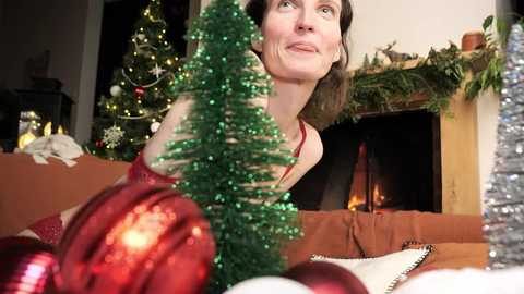 Media: Video of a woman with fair skin and dark hair, wearing a red dress, smiling and looking up. Christmas decorations, including a miniature tree, red ornaments, and a lit fireplace, adorn the background.