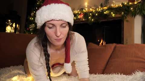 Media: Video of a woman with long, dark hair in a red and white Santa hat, lying on a soft, white shag rug in front of a lit Christmas tree and fireplace.