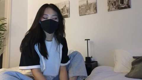 Media: Video of an Asian woman with long black hair, wearing a black face mask, white t-shirt, and grey pajama pants, sitting on a bed, surrounded by black-and-white cityscape videos on a white wall.