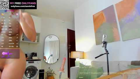 Media: A video of a modern living room with a nude woman standing, viewed from the side. The room features minimalist decor, including a grey couch, a lamp, and abstract art on the walls.