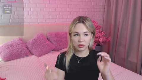 Media: Video of a blonde, fair-skinned woman with light makeup, wearing a black shirt, in a pink-themed room with brick walls, pink pillows, and floral decor.