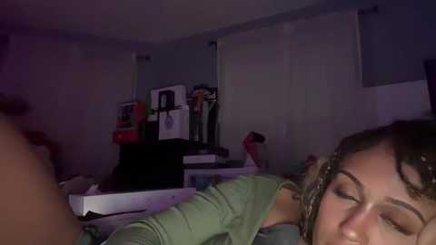 Media: Video of a young woman with light brown hair and closed eyes, resting on a bed with a green shirt, in a dimly lit room with a cluttered desk and white curtains.