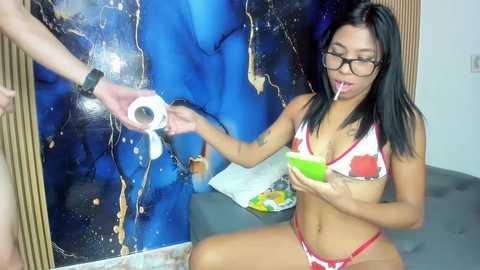 Media: Video of a slender Asian woman with black hair, glasses, and a red bikini, drinking from a green cup, while a hand rolls a condom.