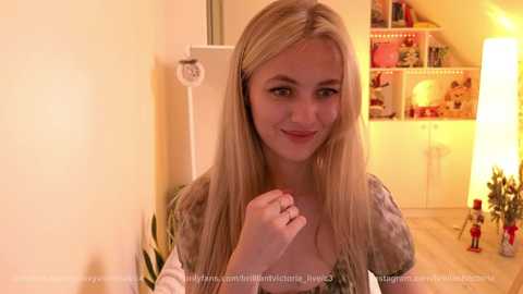 Media: Video of a smiling, blonde woman with straight hair, wearing a tie-dye shirt, standing indoors near shelves filled with toys and a lit lamp.