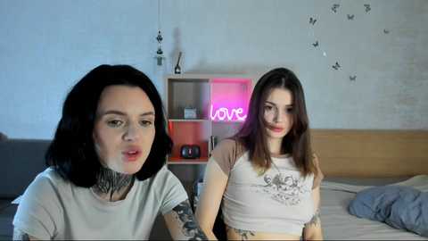 Media: Video of two women with pale skin and dark hair, tattoos, sitting on a bed in a dimly lit room with a neon \"LOVE\" sign and butterfly wall art.