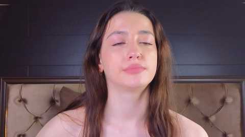 Media: Video of a young woman with long brown hair, fair skin, and closed eyes, wearing a strapless top, standing in front of a dark, tufted headboard.